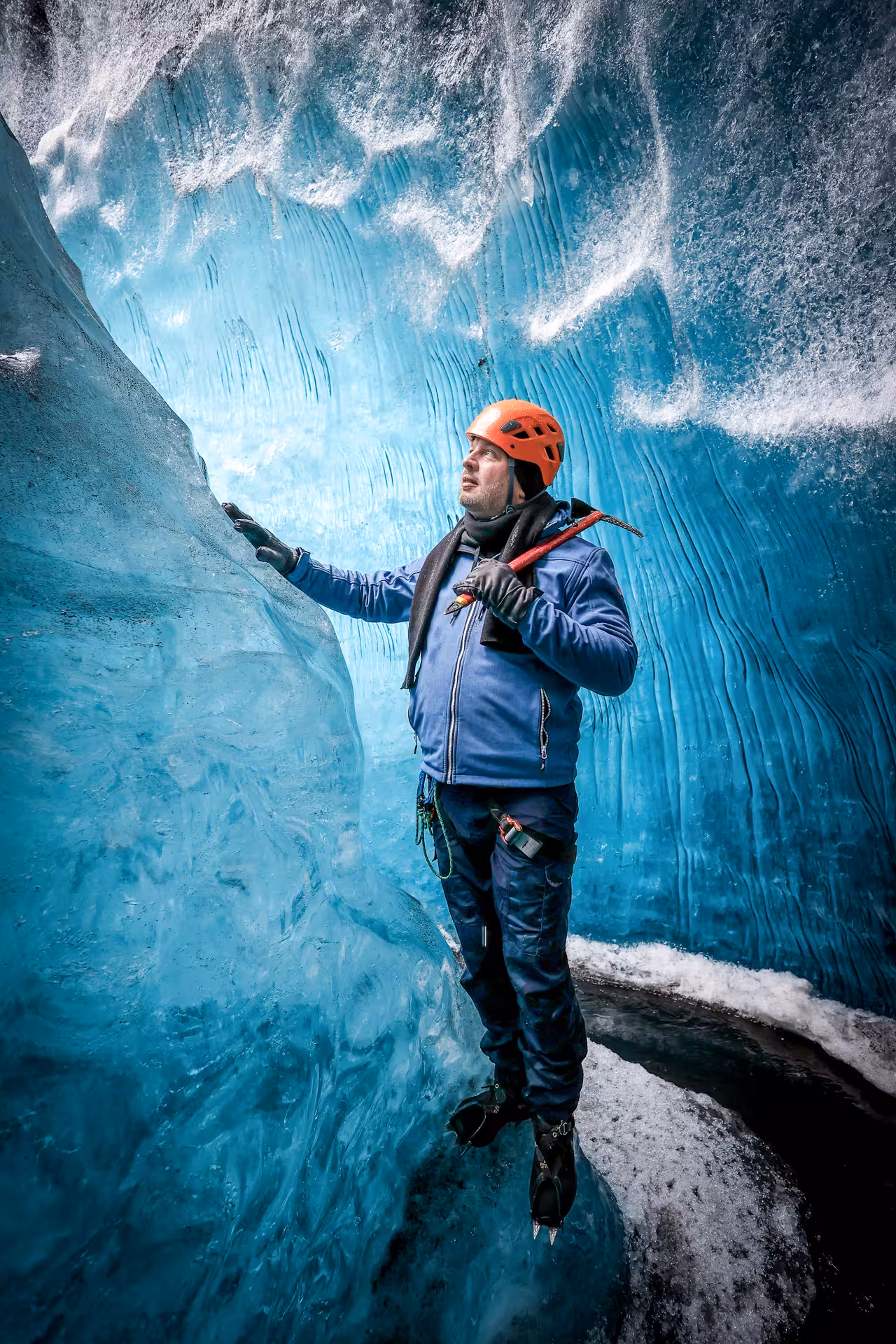 A hiker with red helmet and ice axe explores stunning blue ice cave walls on Falljokull glacier guided tour Iceland