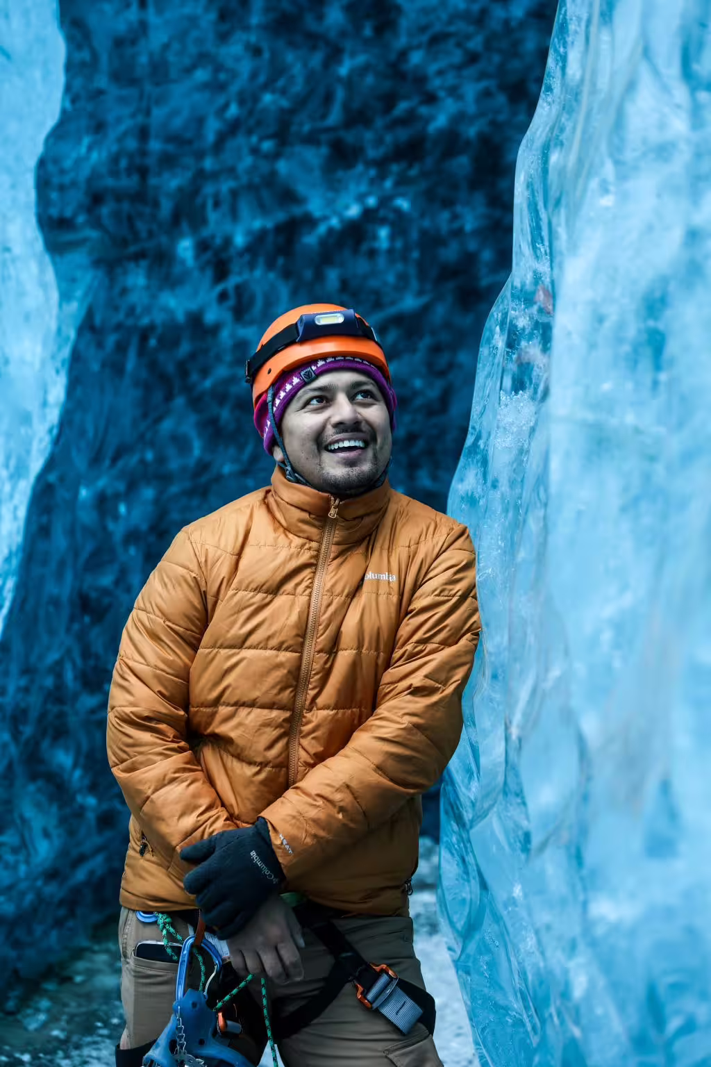 Smiling adventurer in orange helmet and puffer jacket poses inside a stunning blue glacier ice cave tour