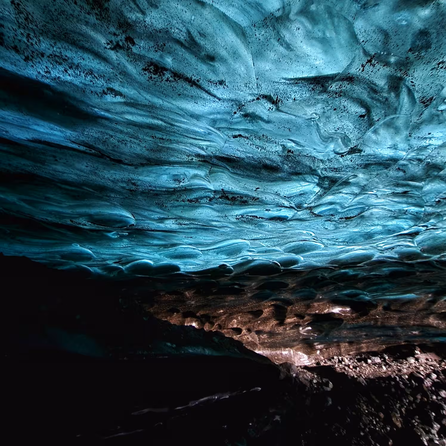 Glowing turquoise blue ice cave ceiling with layered glacier formations and dark rocky floor in Iceland