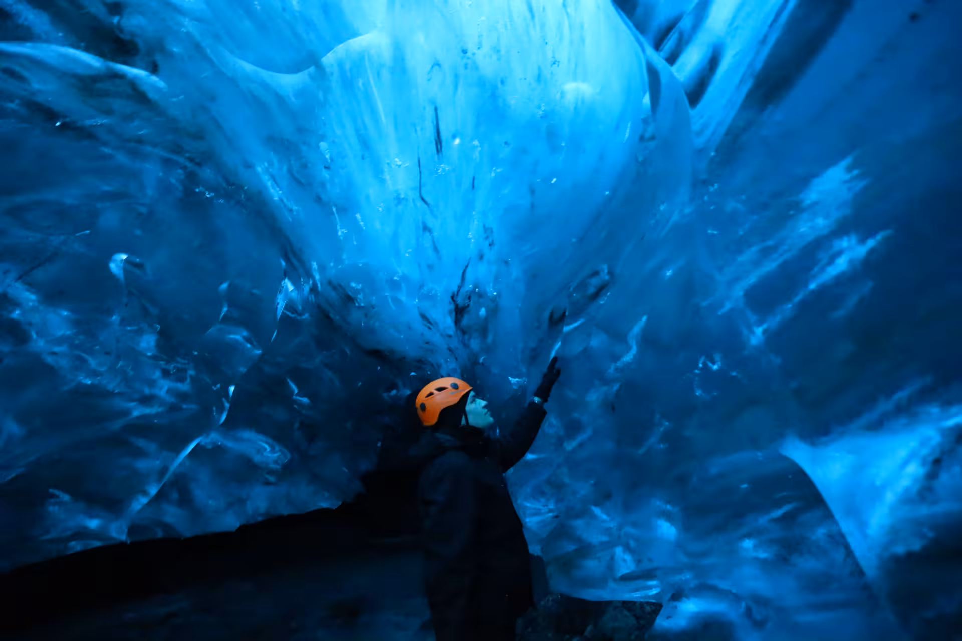 Explorer in orange helmet touching glowing blue ice cave walls during glacier tour in Iceland