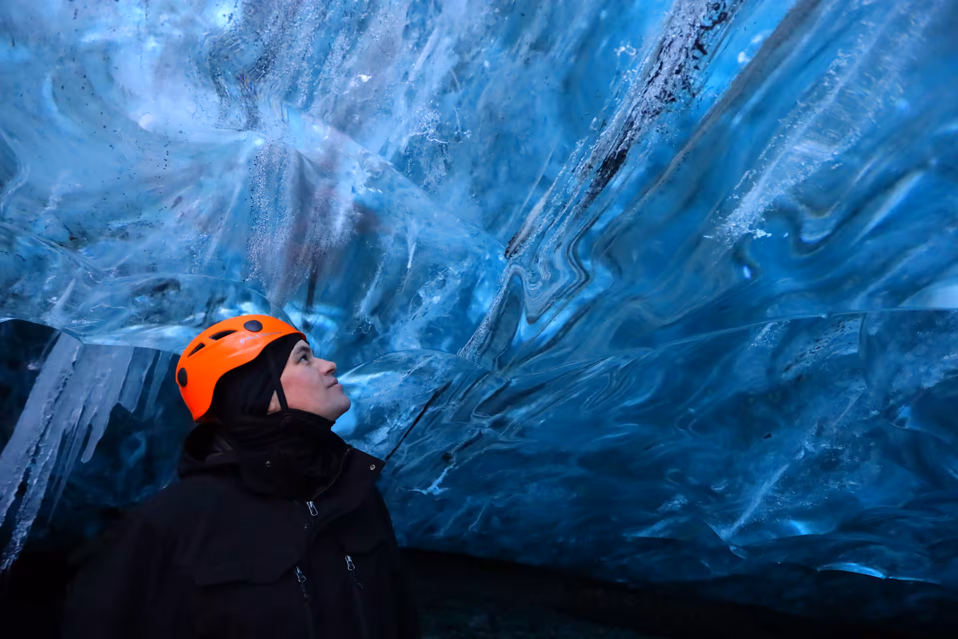 Explorer in orange helmet gazing up at stunning blue glacial ice cave walls in Iceland