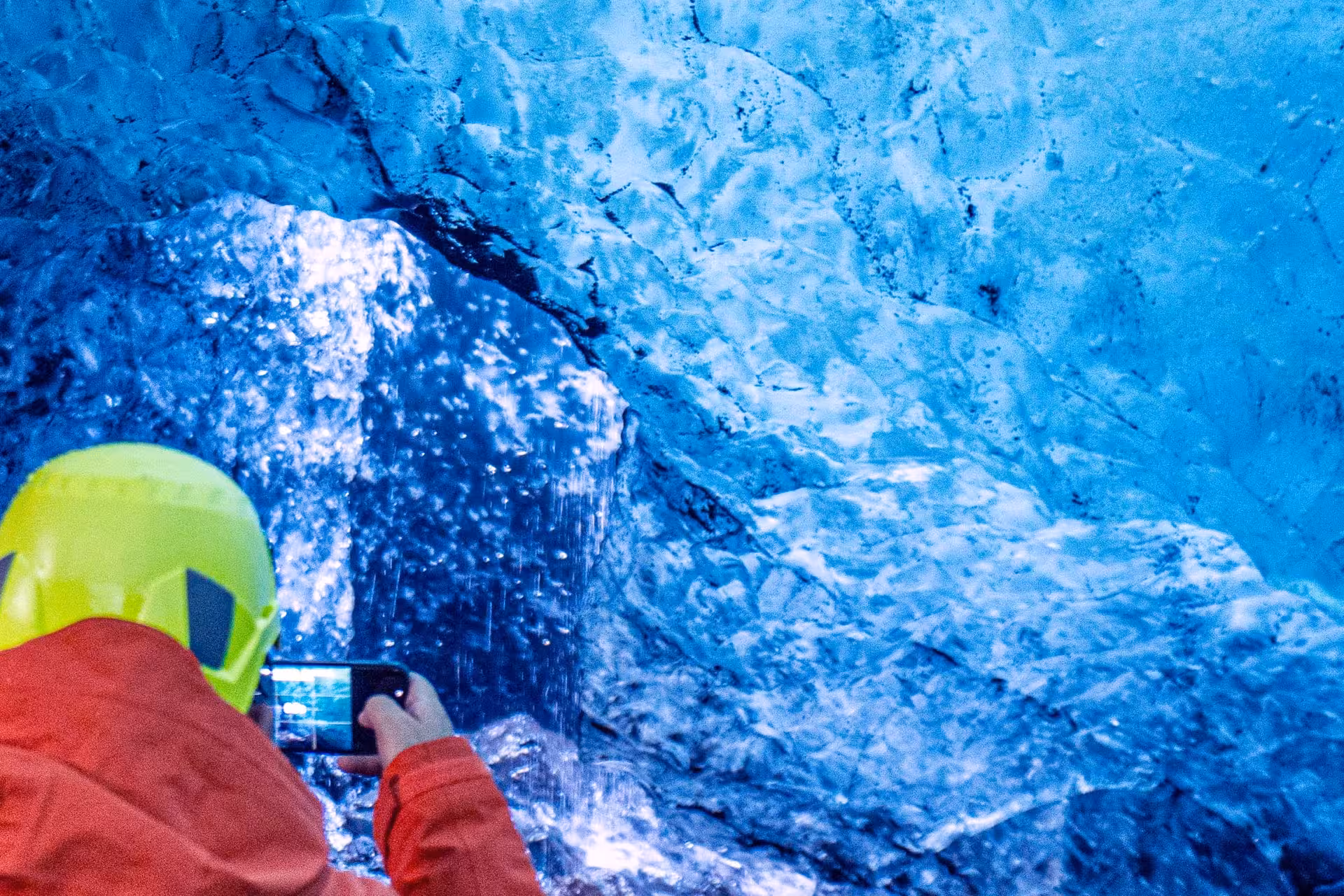 Tourist in red jacket and yellow helmet photographing blue ice cave walls in Iceland