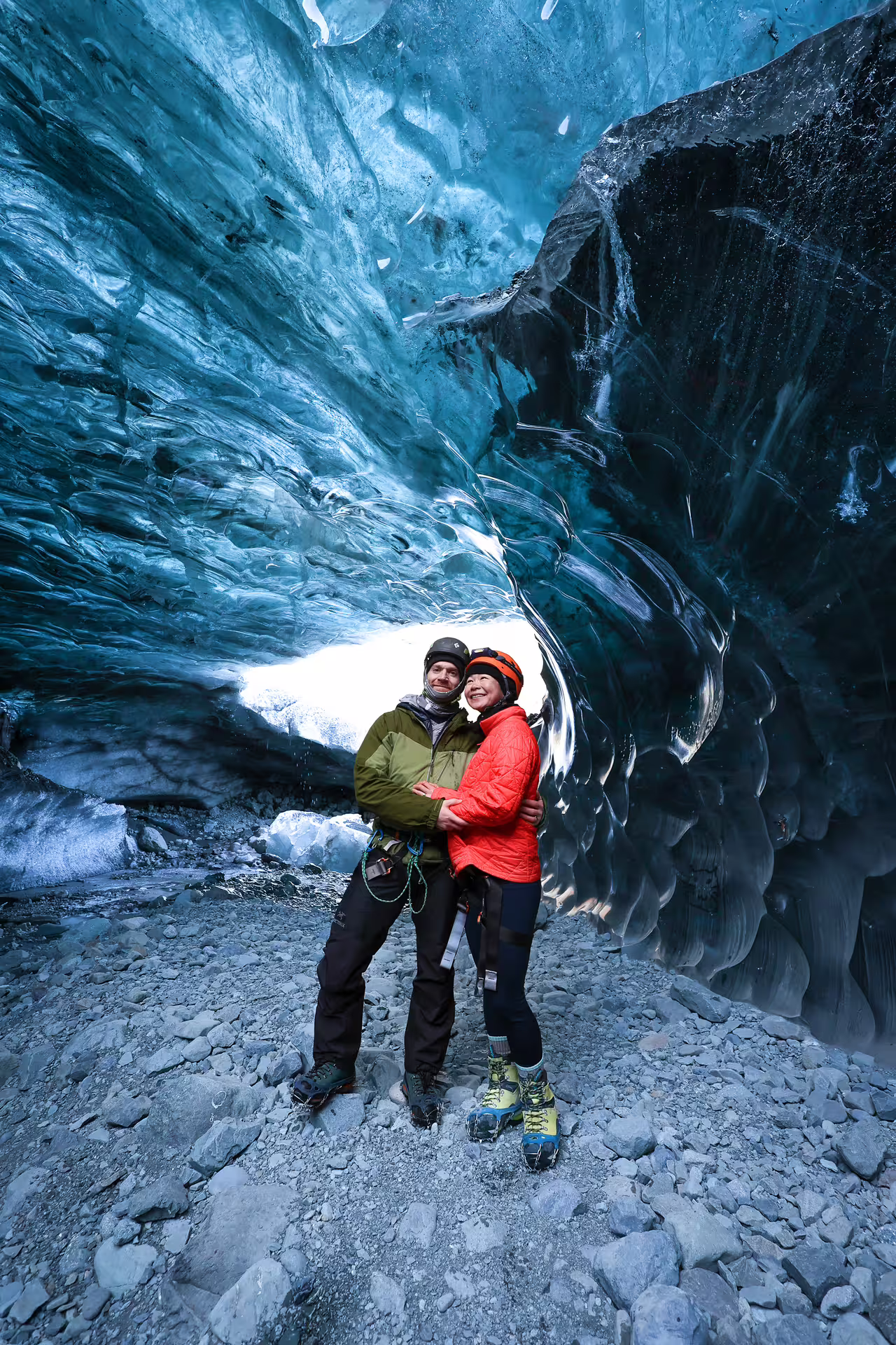 Two adventurers in helmets smiling together inside a vivid blue ice cave on an Iceland glacier hike tour