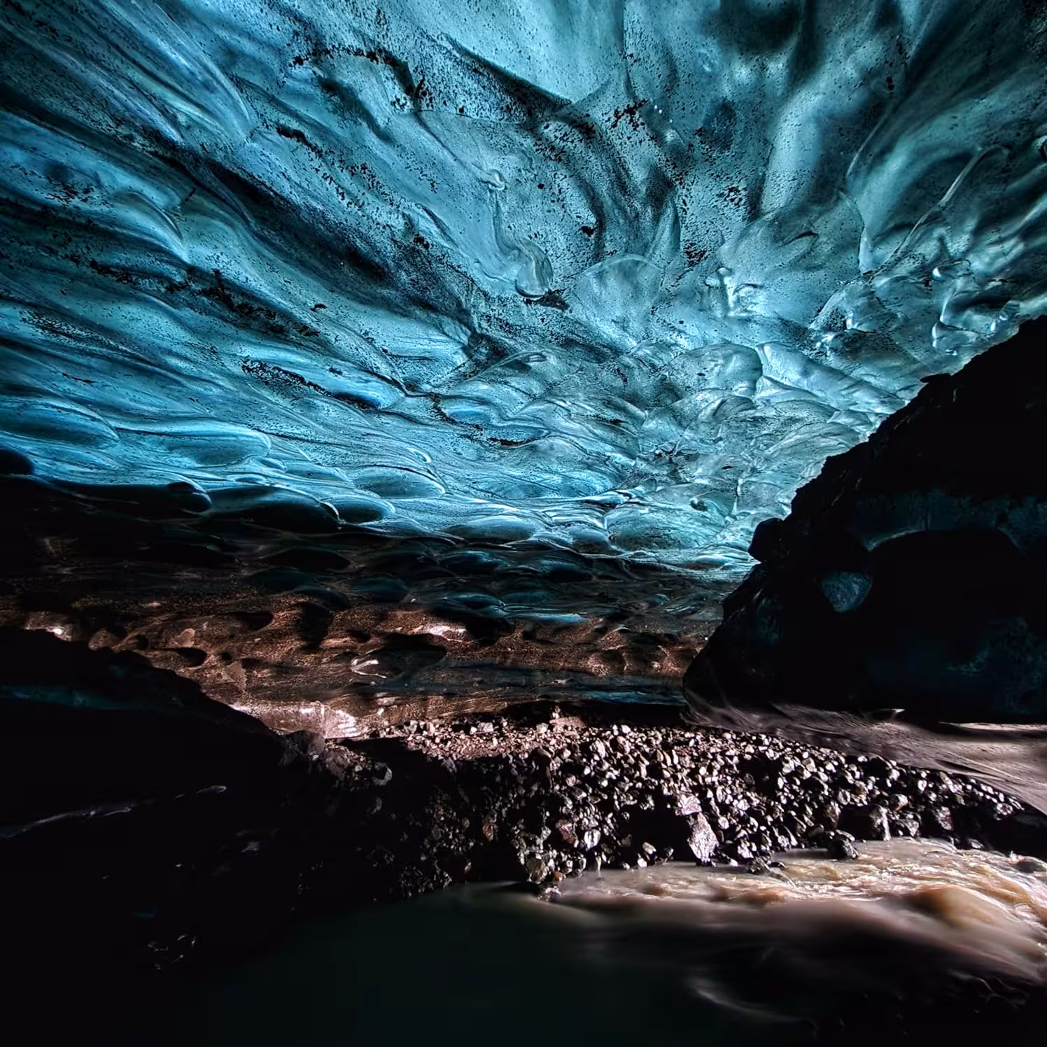 Interior of a blue ice cave in Iceland with glowing turquoise glacier ceiling and rocky stream below