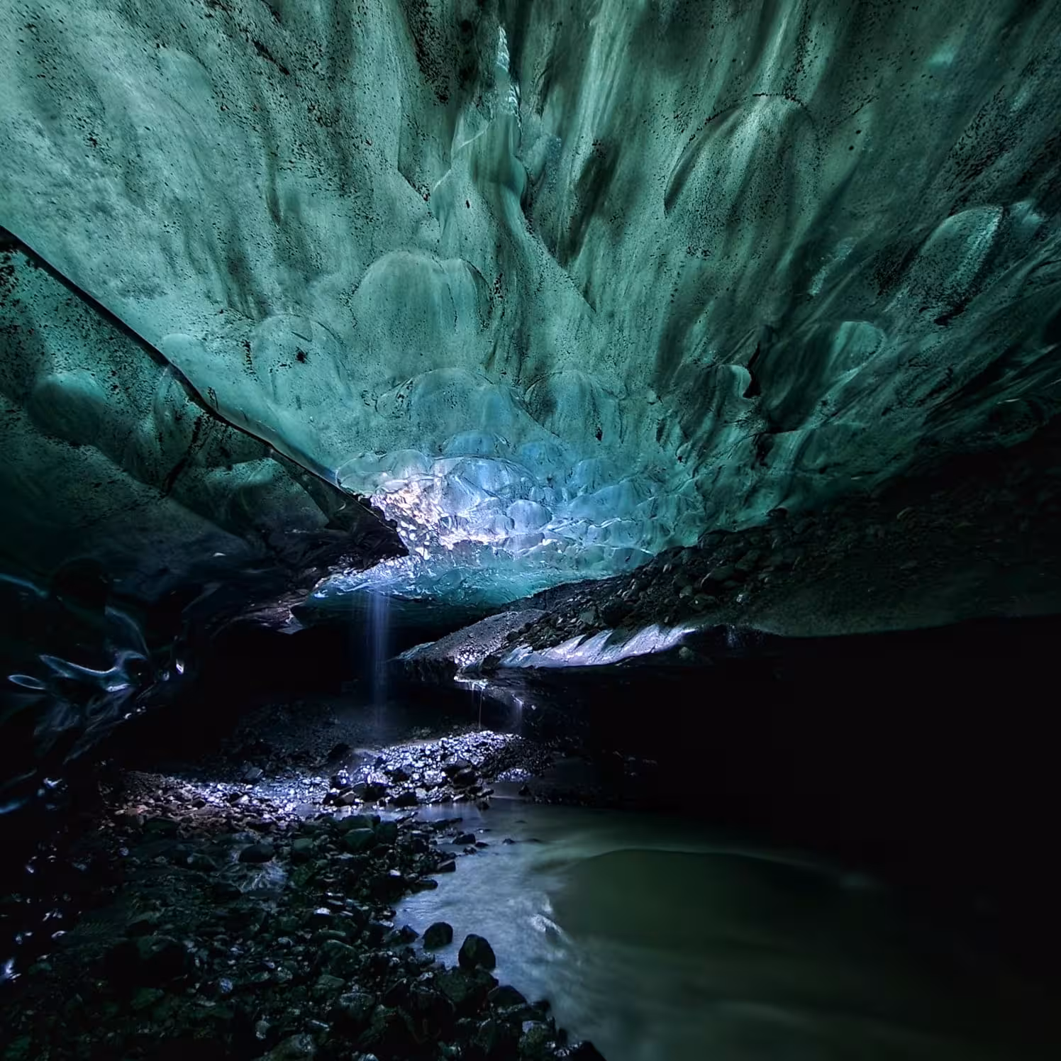 Glowing turquoise blue ice cave ceiling with flowing meltwater stream and rocky floor in Iceland glacier