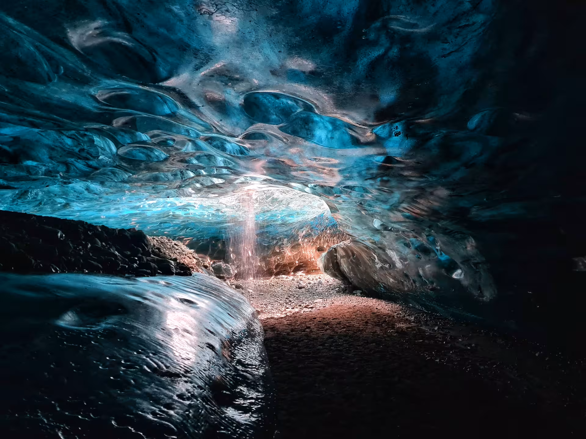 Interior of a blue ice cave in Iceland with glowing turquoise glacier walls and a small waterfall at the entrance