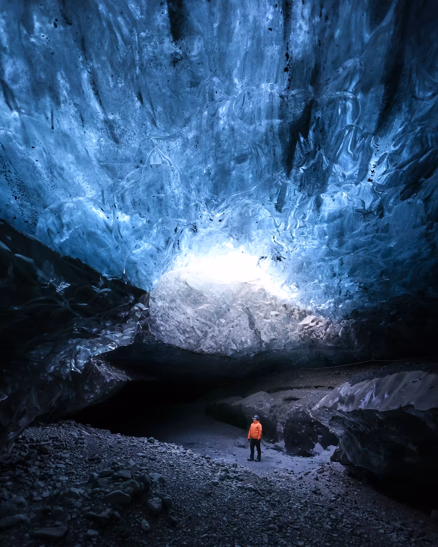 A solo adventurer in an orange jacket stands inside a stunning blue ice cave on a guided glacier tour in Iceland winter.