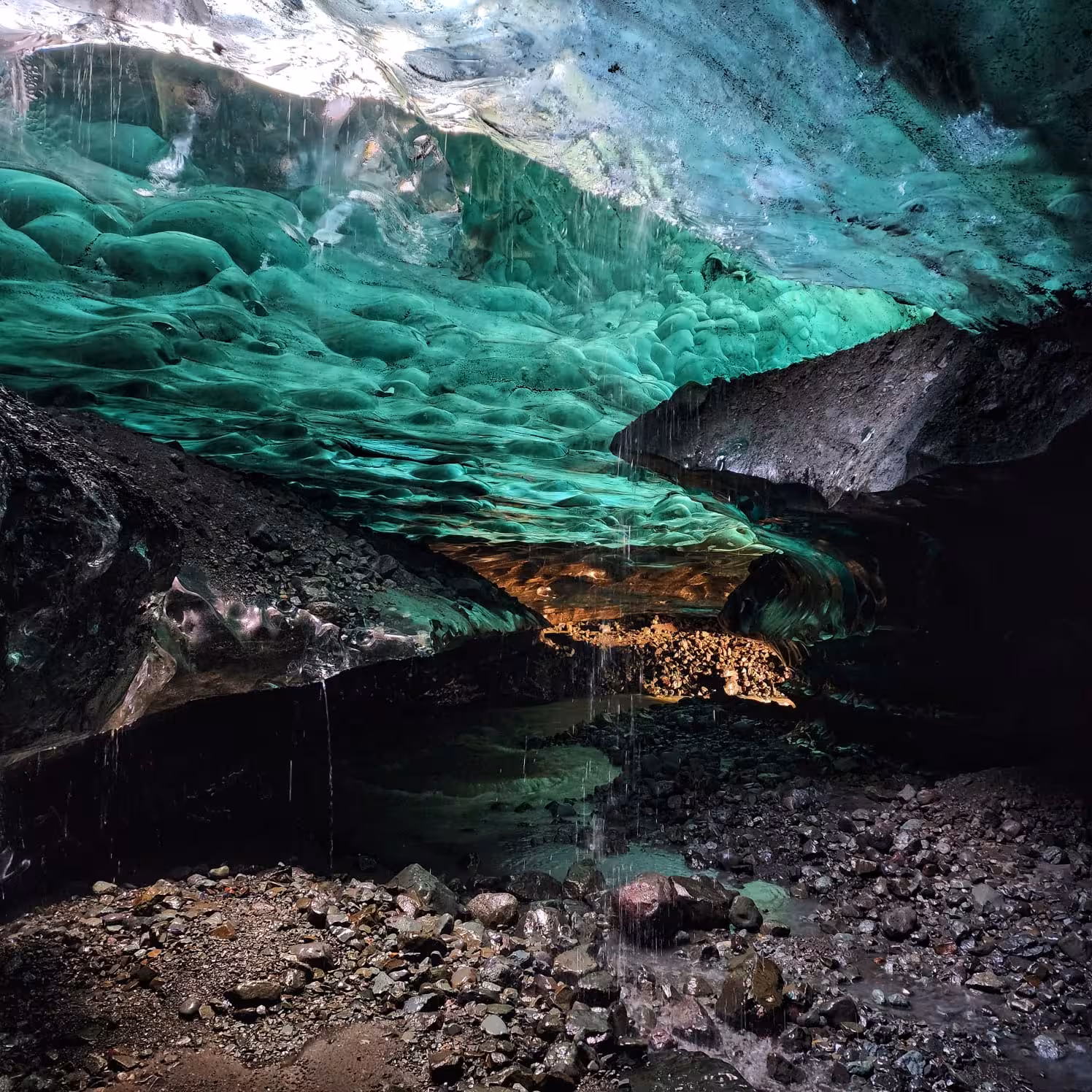 Glowing turquoise blue ice cave ceiling with melting water droplets and rocky floor in Iceland glacier