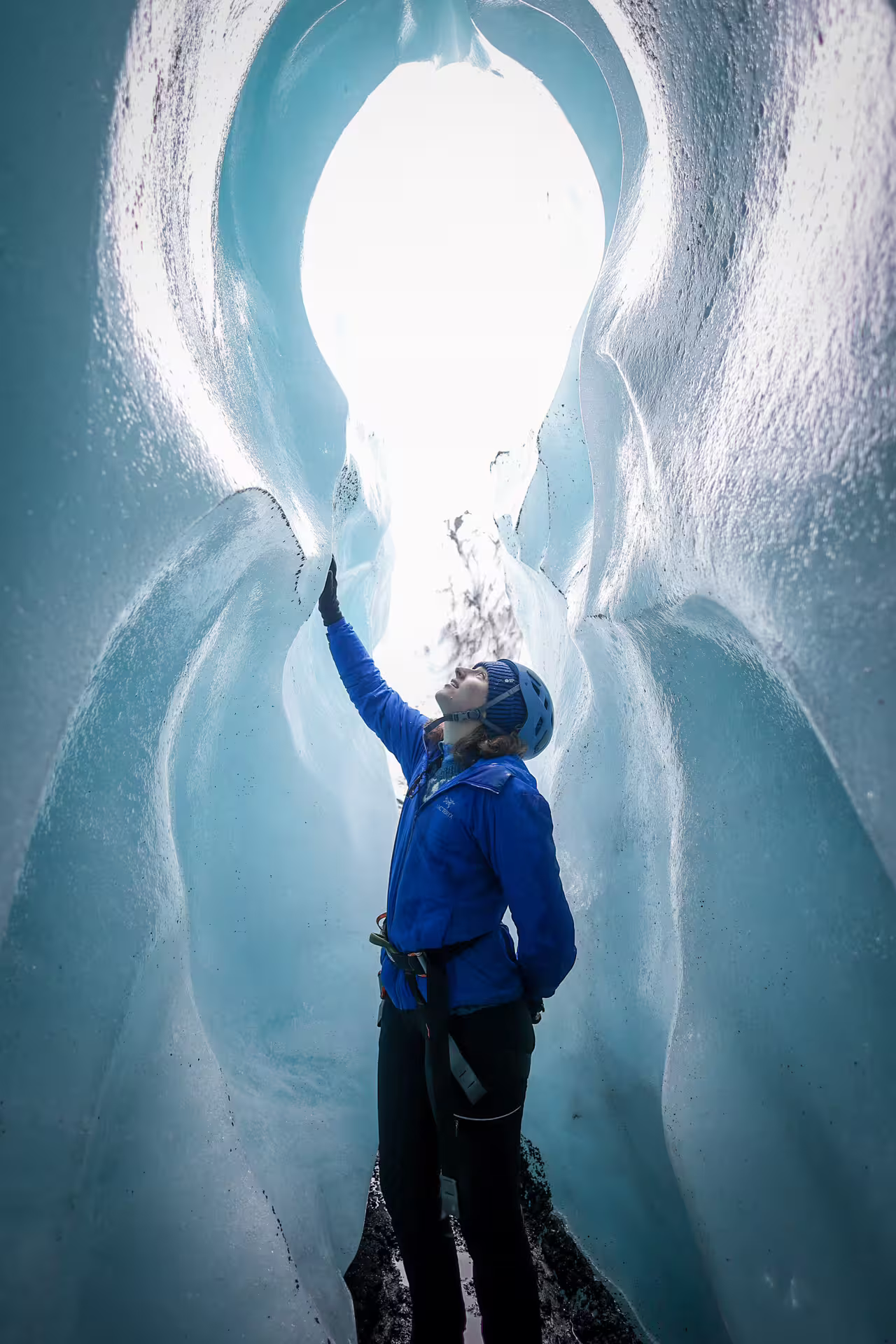A harnessed climber in blue jacket touches glowing blue ice cave walls on Solheimajokull Glacier private ice climbing tour