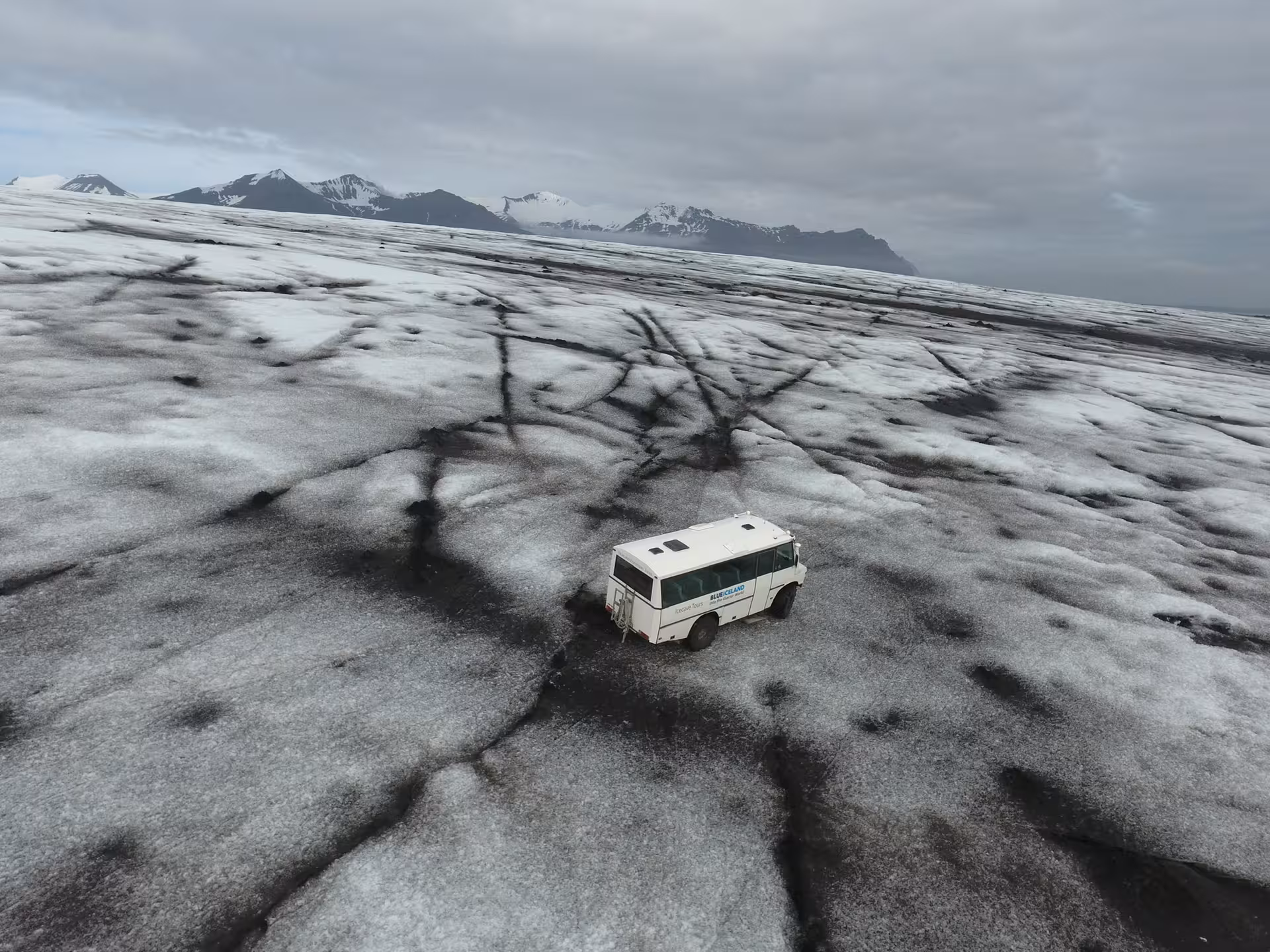 Blue Iceland glacier bus on Vatnajokull ice cap with dark crevasse patterns and snow-capped mountains