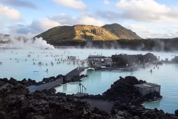 Steaming Blue Lagoon geothermal pool with bathers surrounded by black lava rocks and mountain backdrop in Iceland