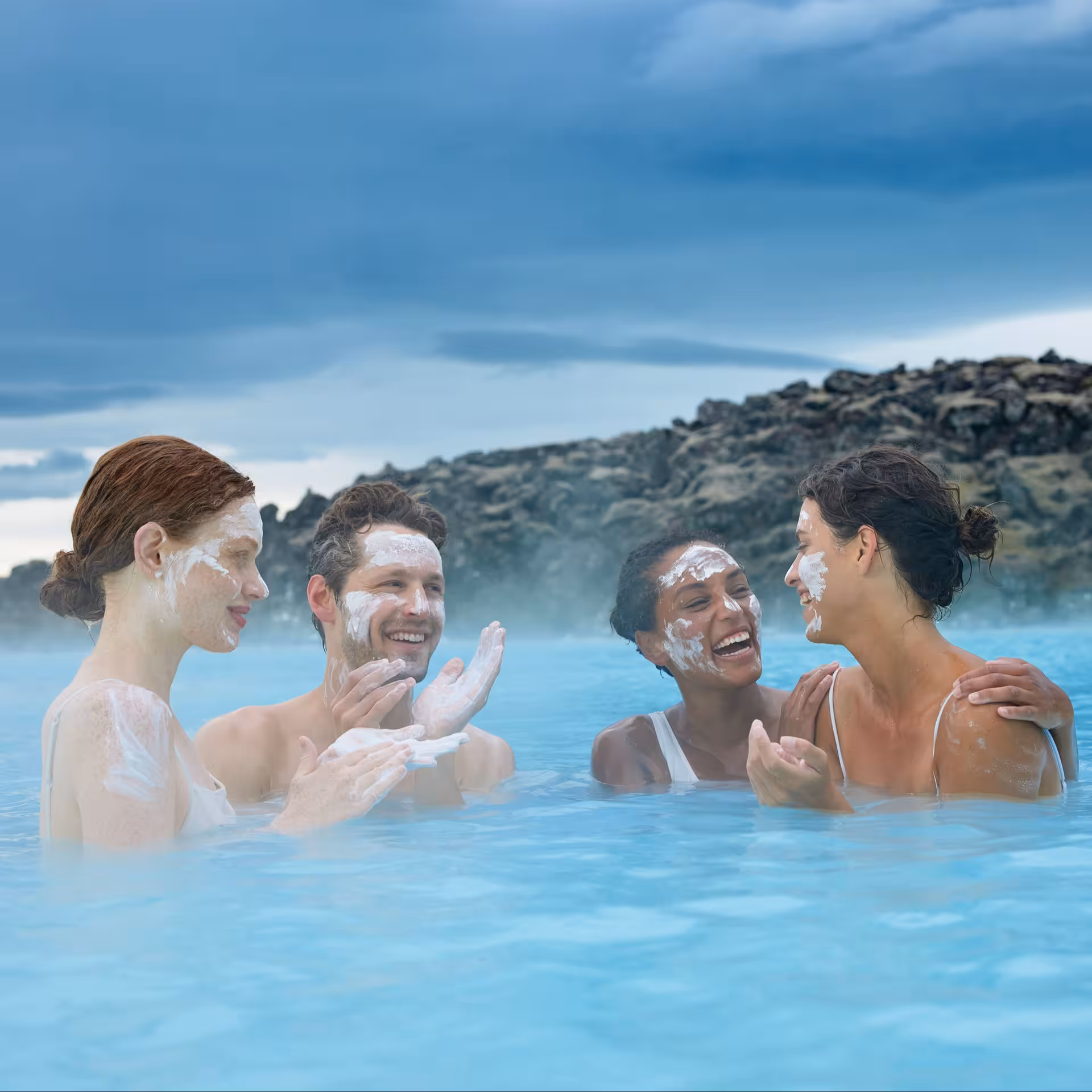Four friends applying silica mud masks while relaxing in Blue Lagoon geothermal hot spring, Iceland