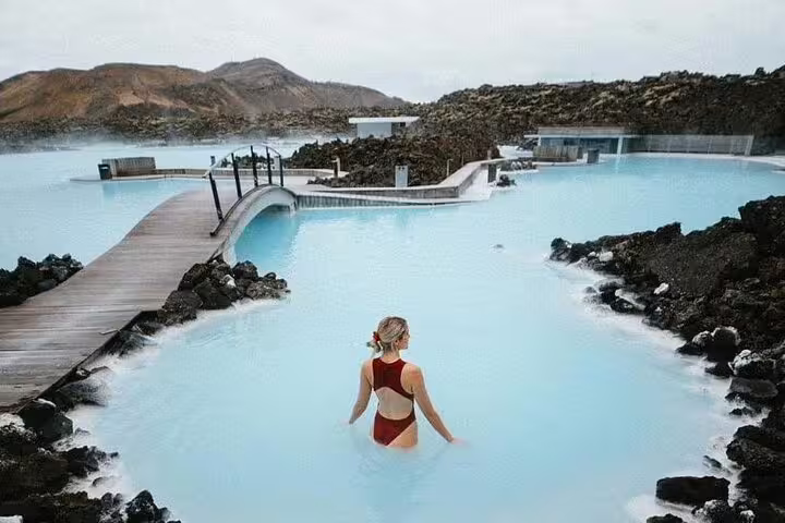Woman in red swimsuit wading in Blue Lagoon geothermal spa with volcanic rocks and boardwalk, Iceland