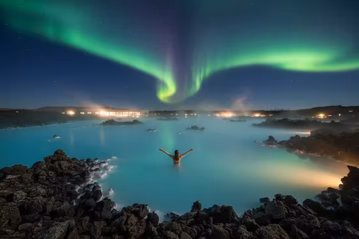 Person soaking in Blue Lagoon geothermal pool under vibrant green aurora borealis in Iceland at night