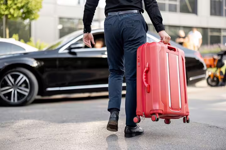 Man in black shirt carrying red suitcase walking toward luxury black sedan for airport transfer