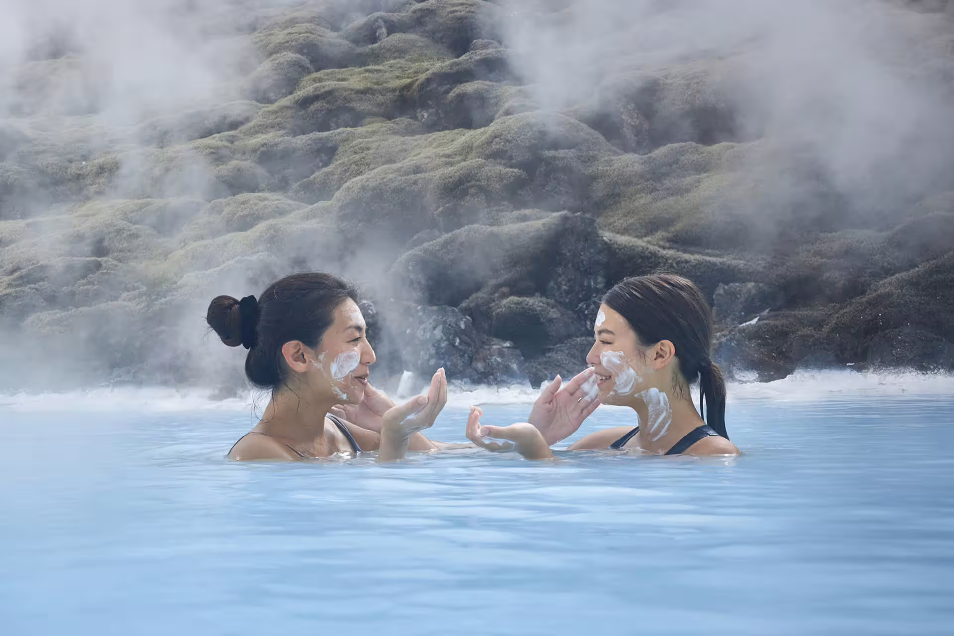 Two women applying white silica mud masks on their faces while soaking in the steaming blue waters of Blue Lagoon