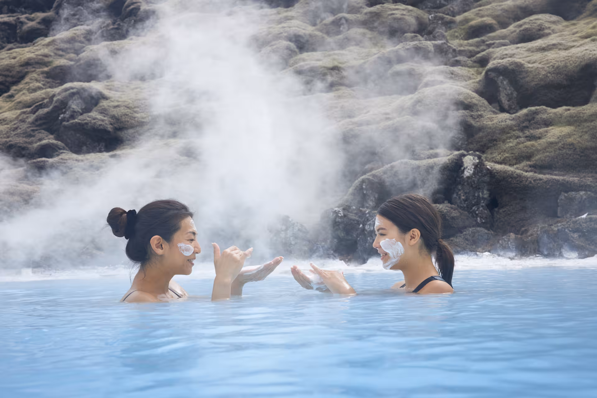 Two people enjoying a relaxing soak in the steaming Blue Lagoon with mud masks, surrounded by rocky landscapes.