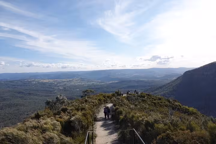 Hikers walking along a scenic path overlooking expansive Blue Mountains landscape under a clear blue sky.