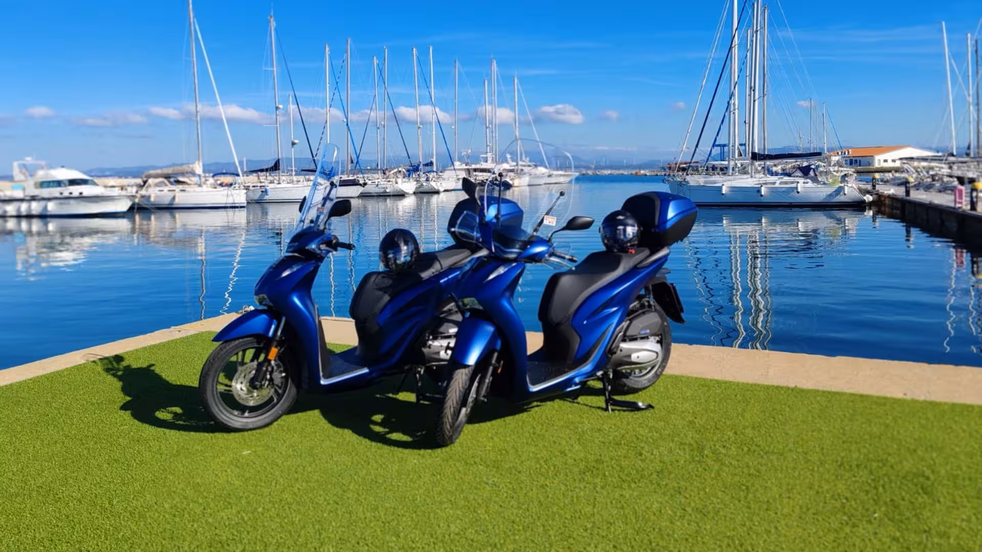 Two blue scooters parked on green grass near a marina in Calasetta, perfect for exploring Sardinia's scenic coast.