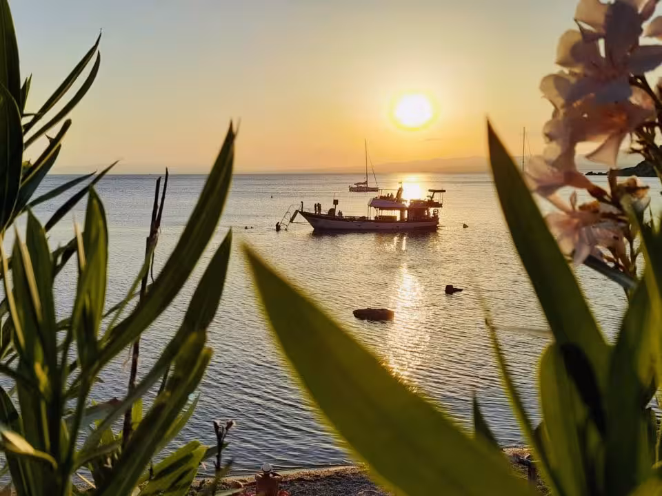 A boat sails at sunset in the serene Capo Rizzuto marine protected area, framed by lush greenery.