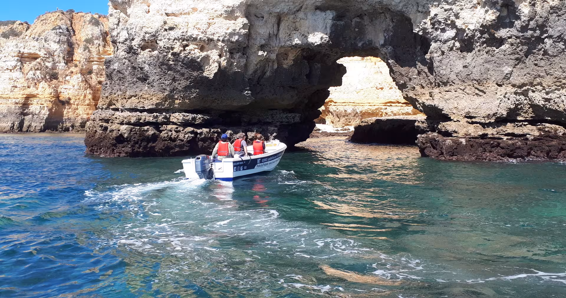 Small boat tour approaches a natural rock arch at Ponta da Piedade caves in Lagos, cruising over clear turquoise water
