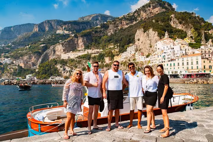 Group of tourists posing by a scenic boat with Amalfi's picturesque cliffs and town in the background.
