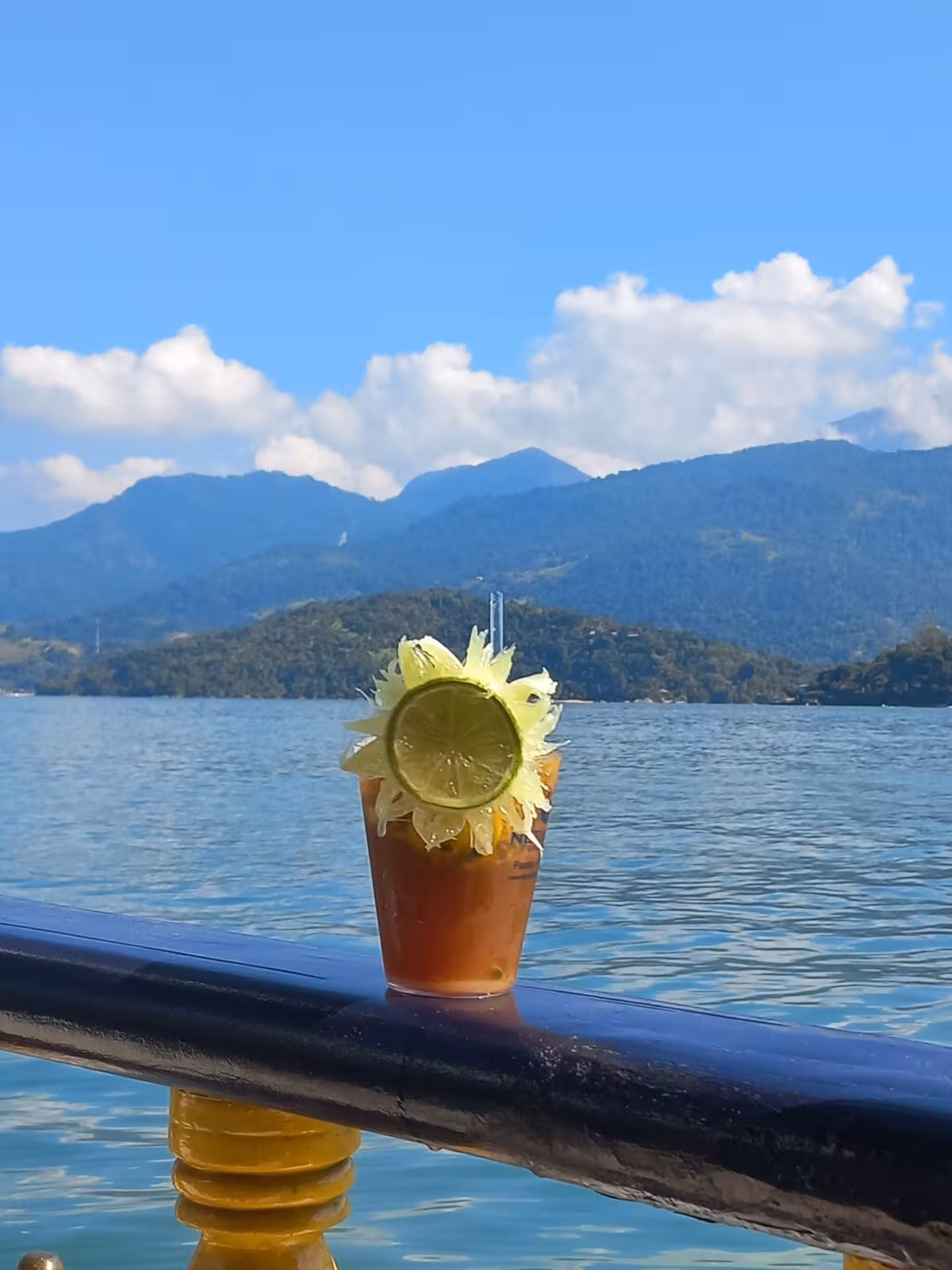 Refreshing caipirinha with lime garnish on a boat railing, set against a stunning mountain and sea backdrop.