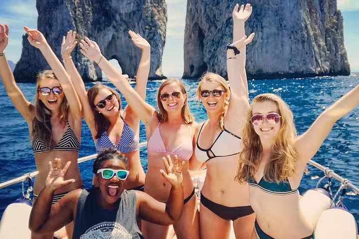 Excited friends on boat trip to Capri Island from Sorrento, smiling with iconic Faraglioni rocks in background.