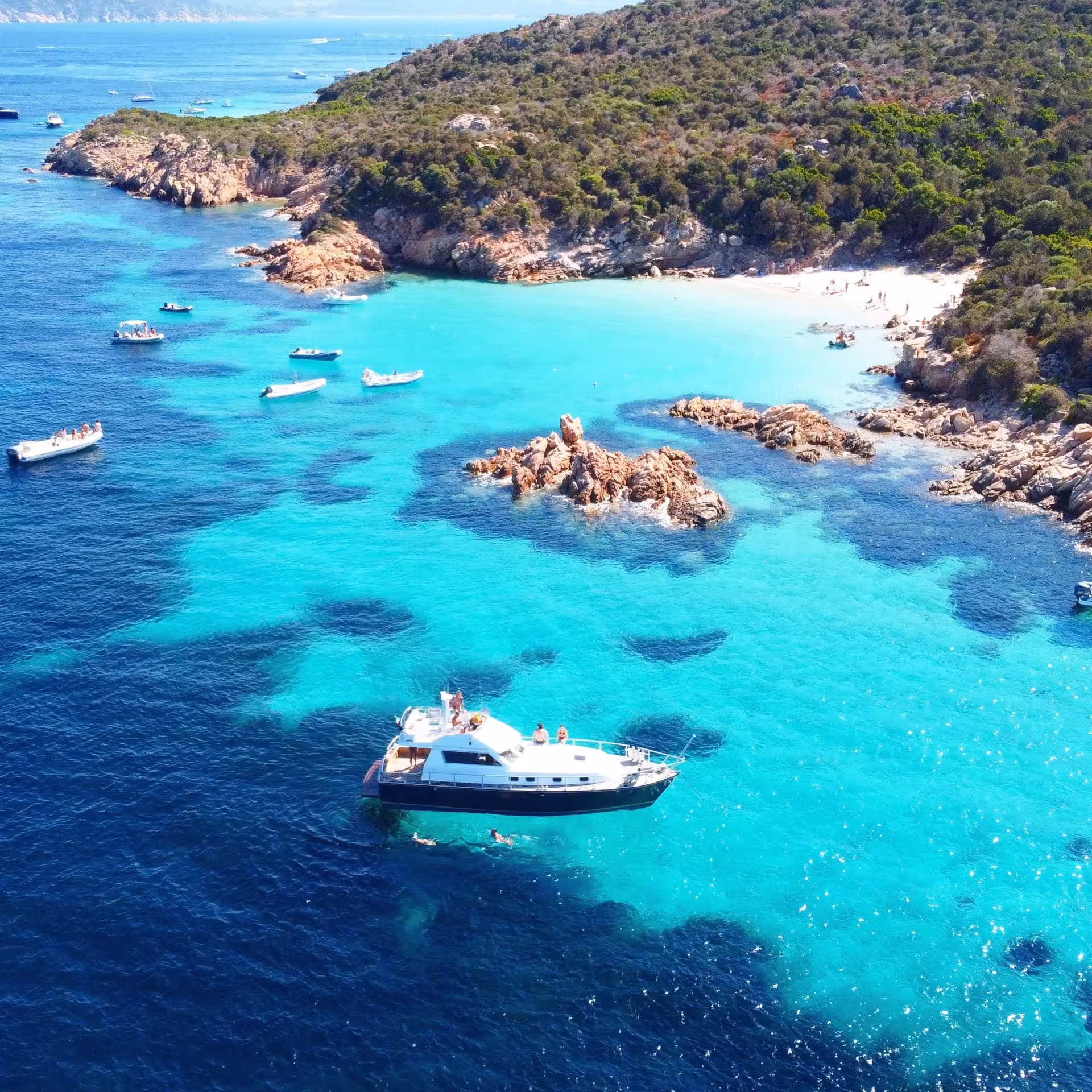 Aerial view of a boat anchored in the turquoise waters of La Maddalena Archipelago near a rocky shoreline.