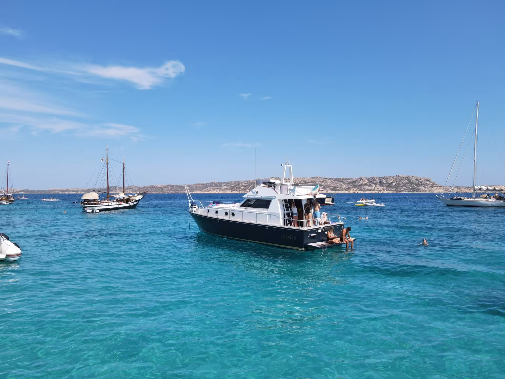Tourists enjoying a boat trip on turquoise waters with multiple sailboats and a scenic backdrop in La Maddalena Archipelago.