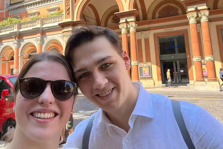 Couple smiling in selfie in front of historic Bologna arcade building on sunny day