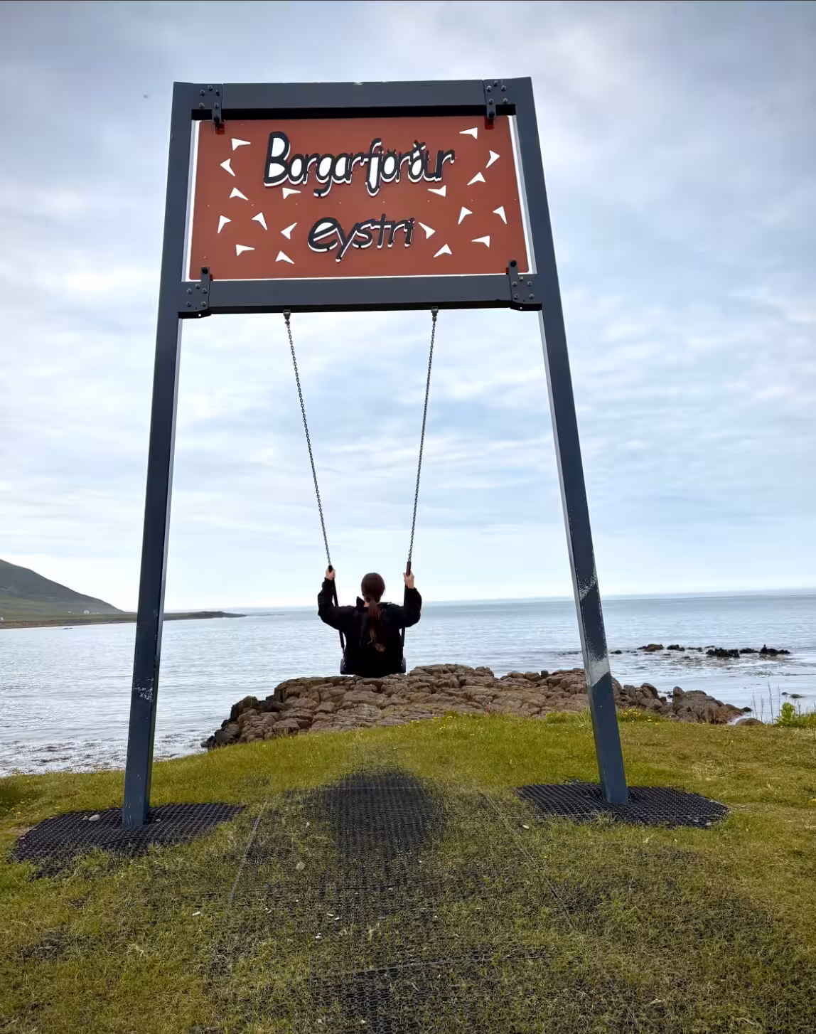 Person sitting on swing at Borgarfjörður Eystri sign overlooking calm Eastfjords coastline in Iceland
