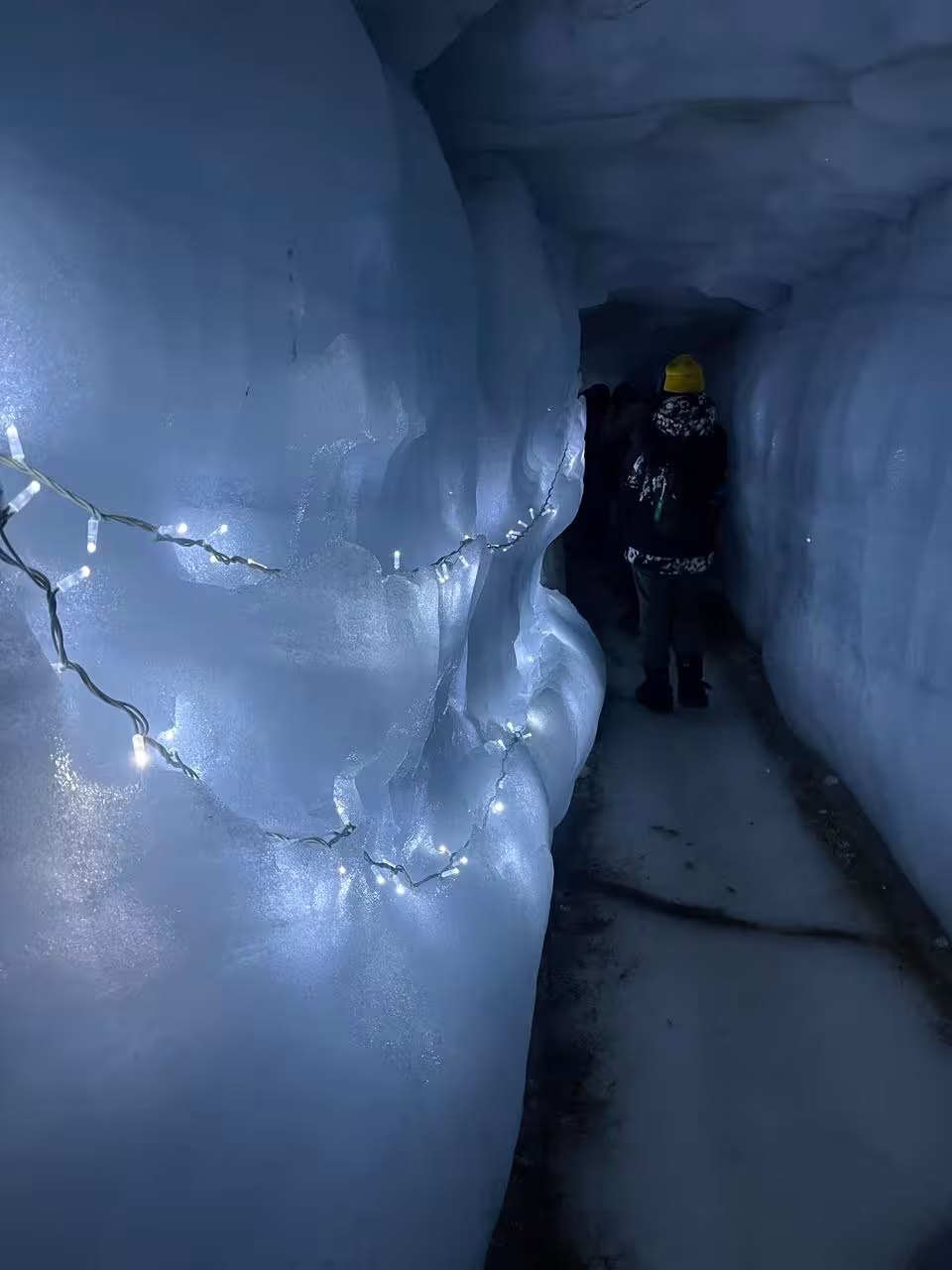 Visitors walking through a narrow illuminated ice cave tunnel with string lights in Borgarfjordur Iceland