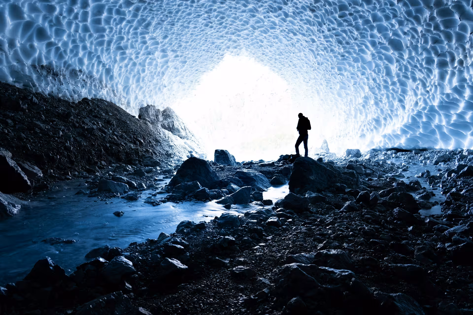 Silhouetted hiker standing inside a glacial ice cave with rocky floor and bright entrance in West Iceland