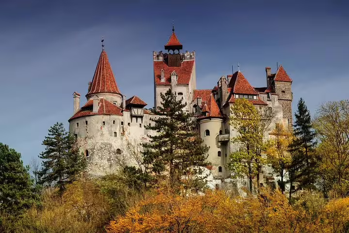 Bran Castle in Transylvania with red-roofed towers surrounded by autumn trees under blue sky