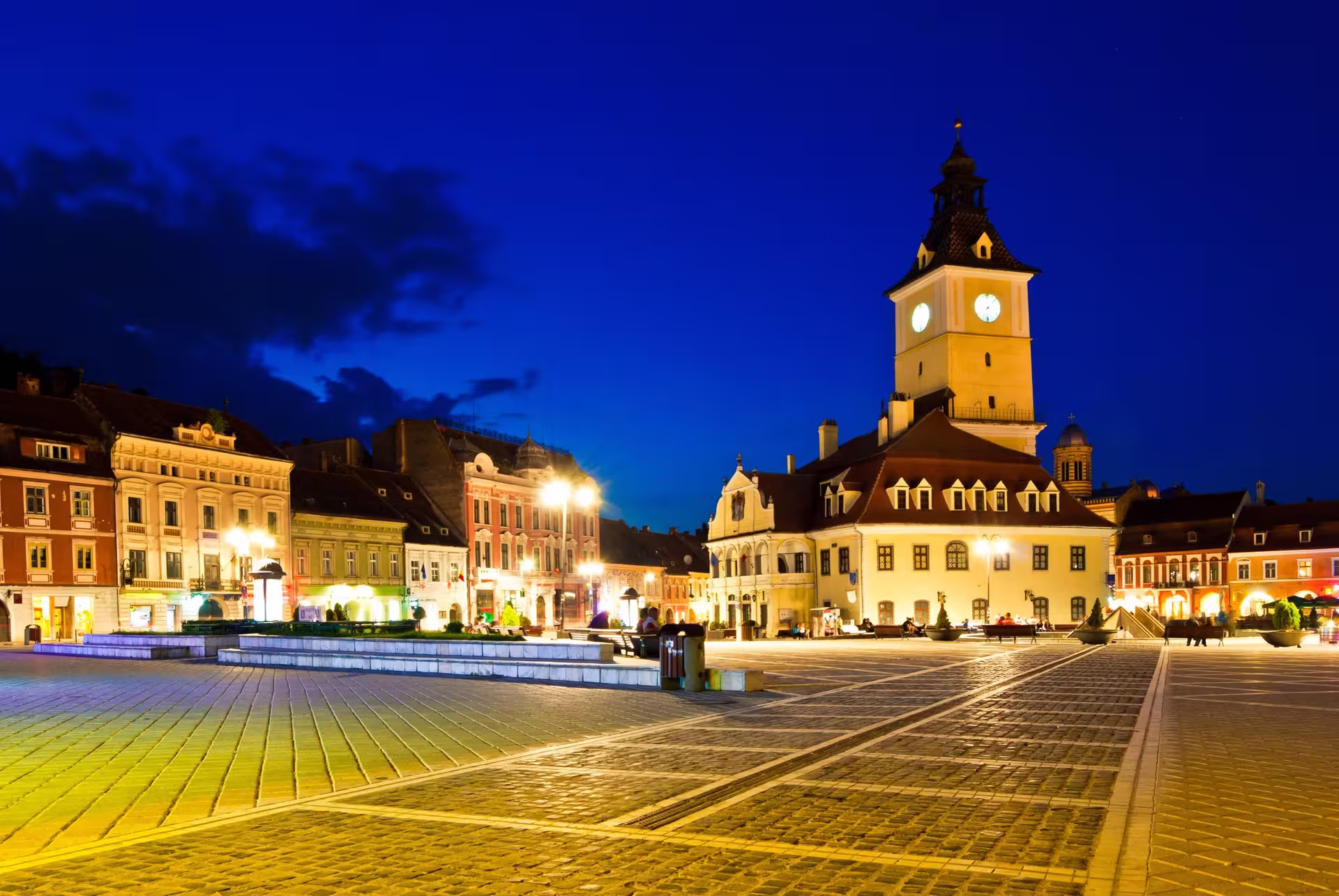 Night view of Brasov's illuminated town square, showcasing historic buildings and vibrant nightlife ambiance.