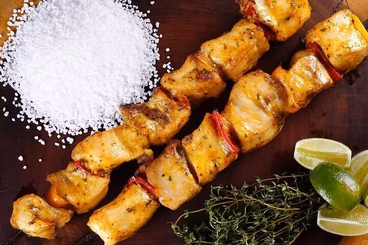Grilled chicken skewers seasoned with herbs and coarse salt, accompanied by fresh limes and thyme on a wooden board.