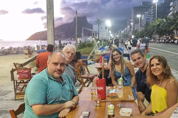 Group enjoying Brazilian cocktails at a beachfront cafe on Ipanema Beach, with a scenic sunset in the background.
