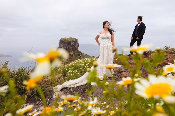 Bride and groom posing on Santorini cliff with wildflowers, captured by a wedding proposal photographer session