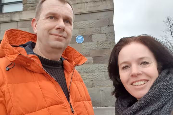 Couple smiling on Bristol waterfront walking tour near stone building with blue heritage plaque