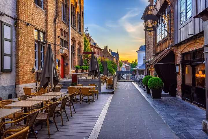 Charming canal-side street in Bruges with outdoor cafe seating, brick buildings, and hotel sign at sunset