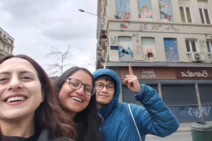 Three smiling tourists taking a selfie in front of a colorful comic strip mural on a Brussels building