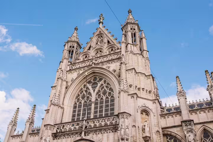 Gothic facade of a Brussels cathedral with ornate stonework, pointed spires, and large arched window against blue sky