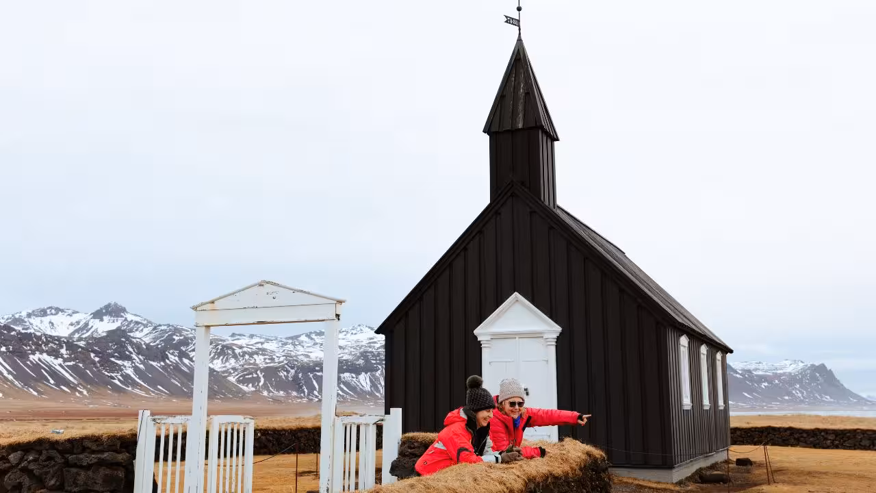 Two tourists in red jackets at the iconic black Búðakirkja church with snow-capped Snæfellsnes mountains behind