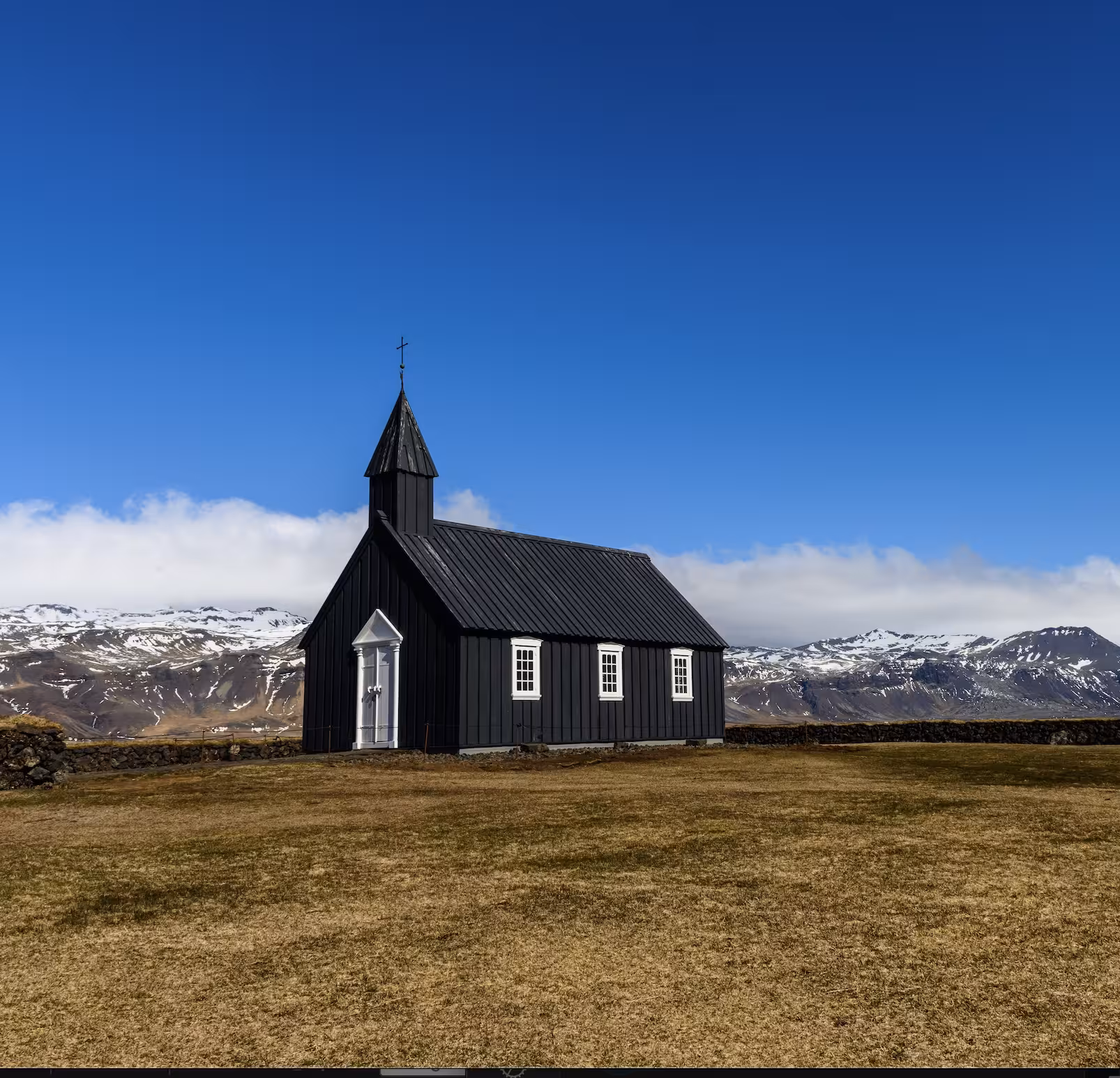 Black Búðakirkja church with white trim and cross against snow-capped Snæfellsnes mountains, Iceland