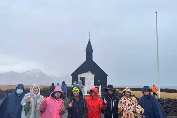 Group of tourists in rain gear posing before Búðakirkja black church on Snæfellsnes Peninsula, Iceland