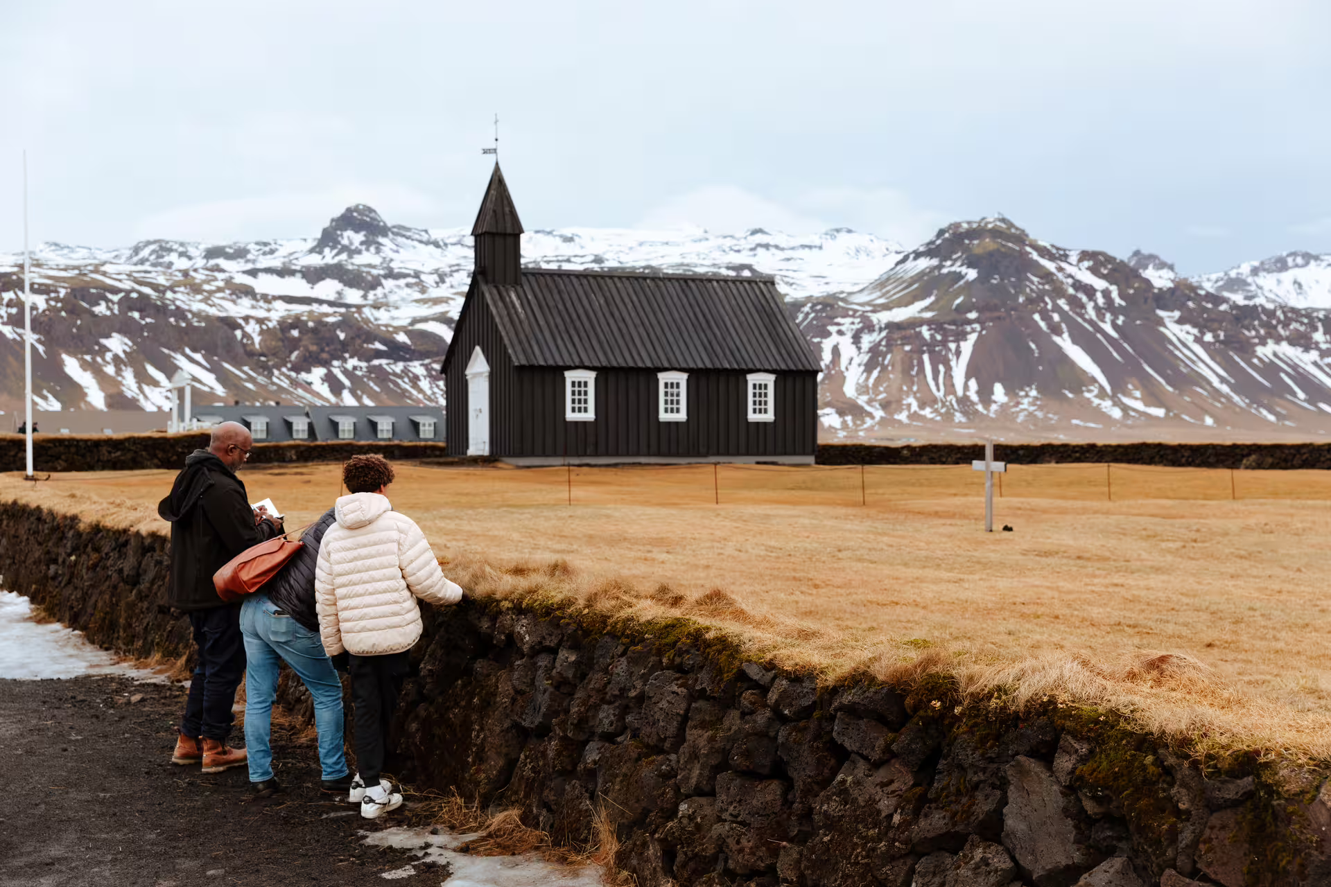 Two tourists overlooking Búðakirkja black church with snow-capped Snæfellsnes mountains in Iceland