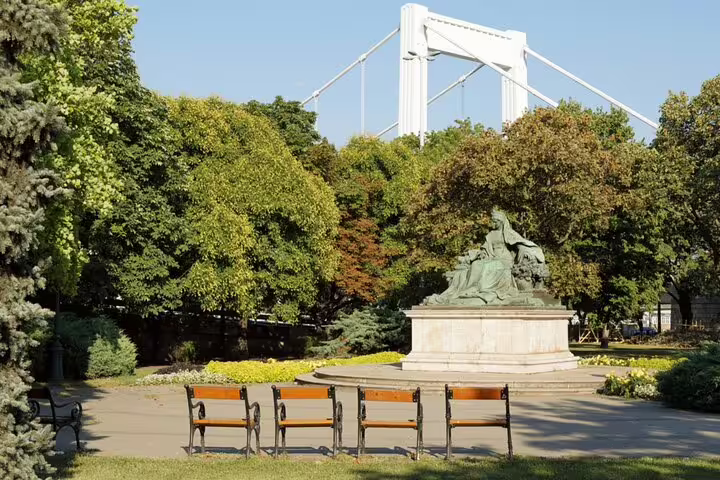 Park benches facing a bronze statue with Elisabeth Bridge visible through trees in Budapest