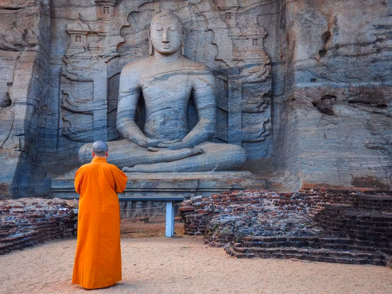 A Buddhist monk in orange robes stands before a large ancient Buddha statue carved into a Sri Lankan rock face.