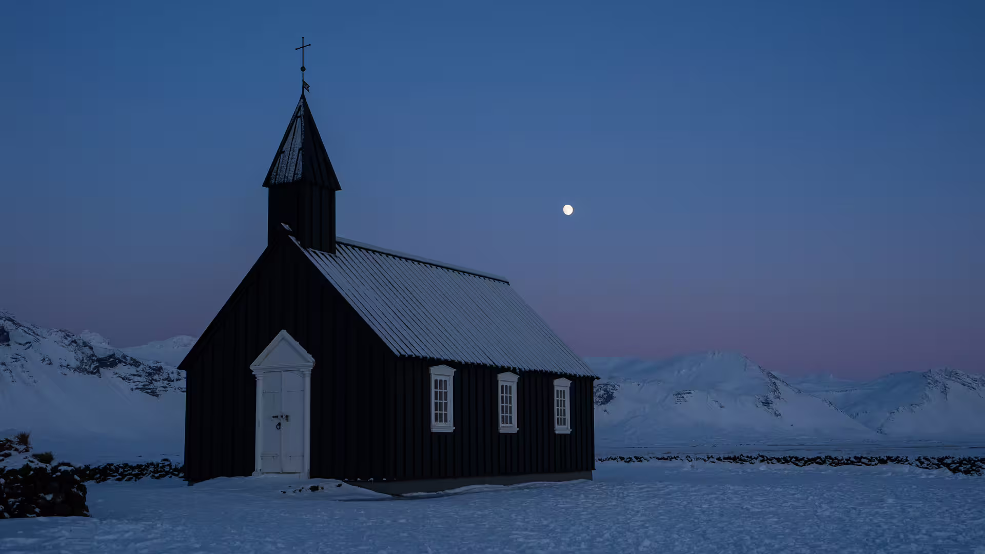 Black church of Budir covered in snow at dusk with moon and snowy mountains, Snaefellsnes Peninsula Iceland